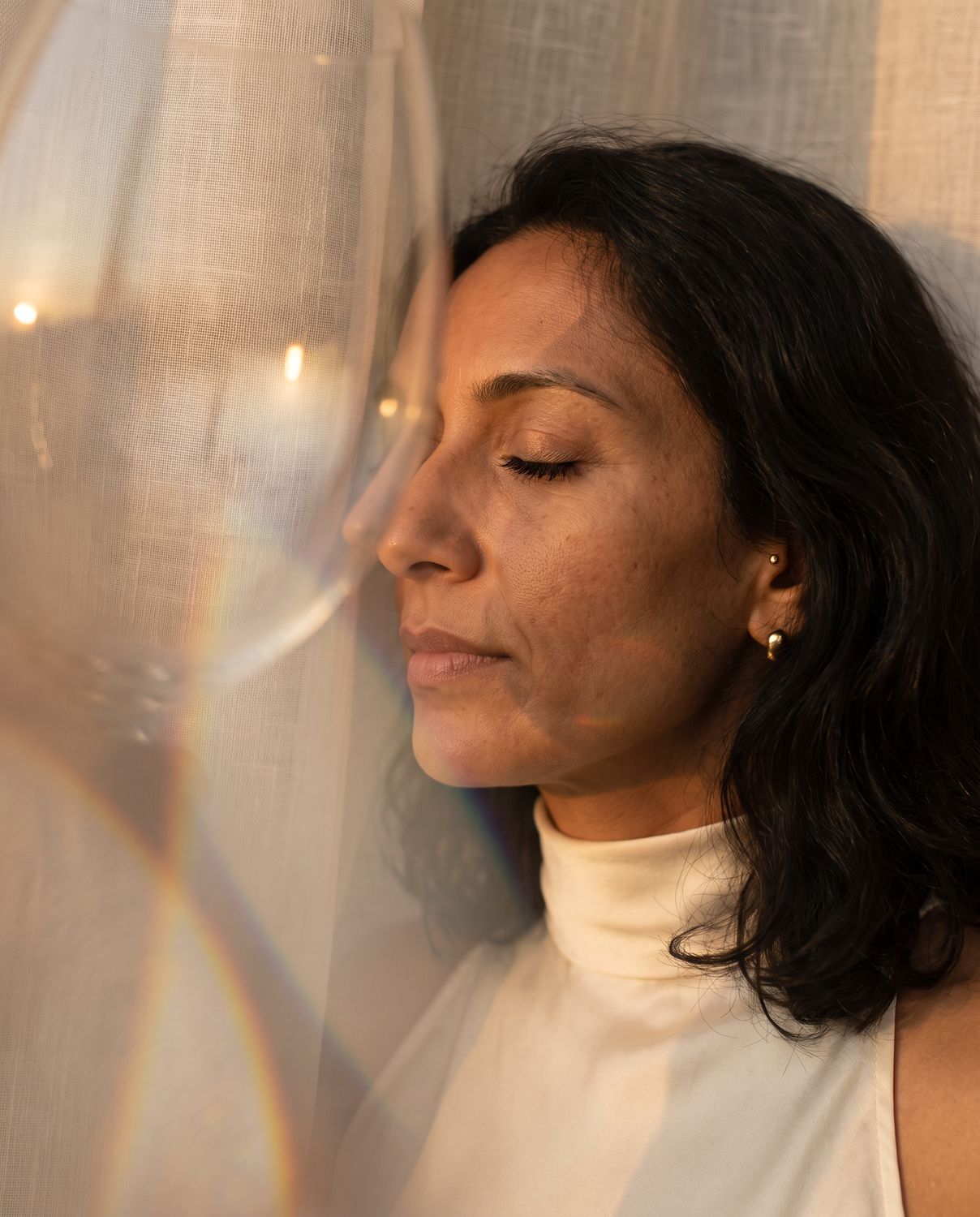 A woman resting her face against a sheer curtain in golden light